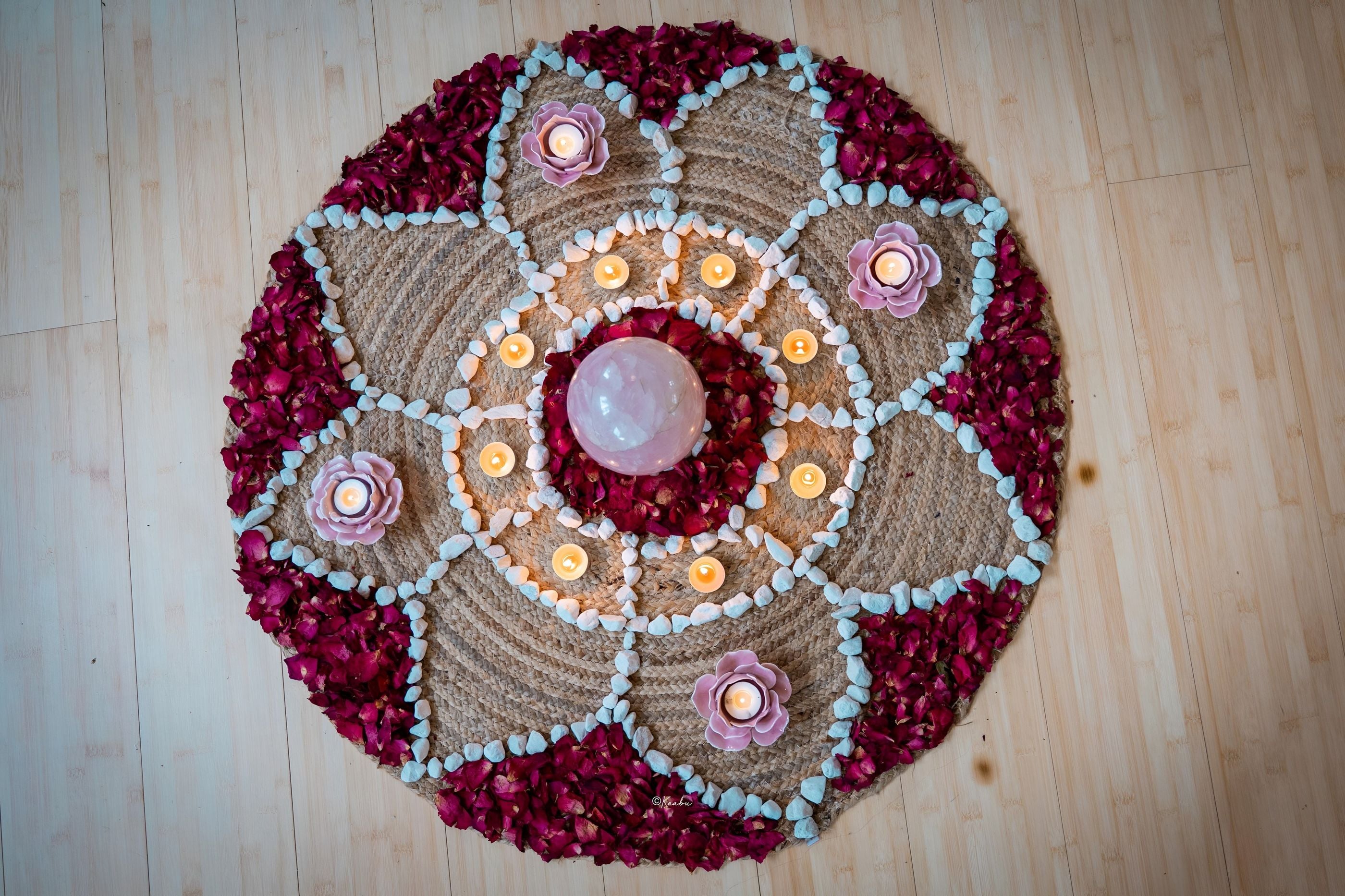 Vu de dessus, un mandala de pétales de rose, de galets blancs et de bougies autour d’une sphère de quartz rose