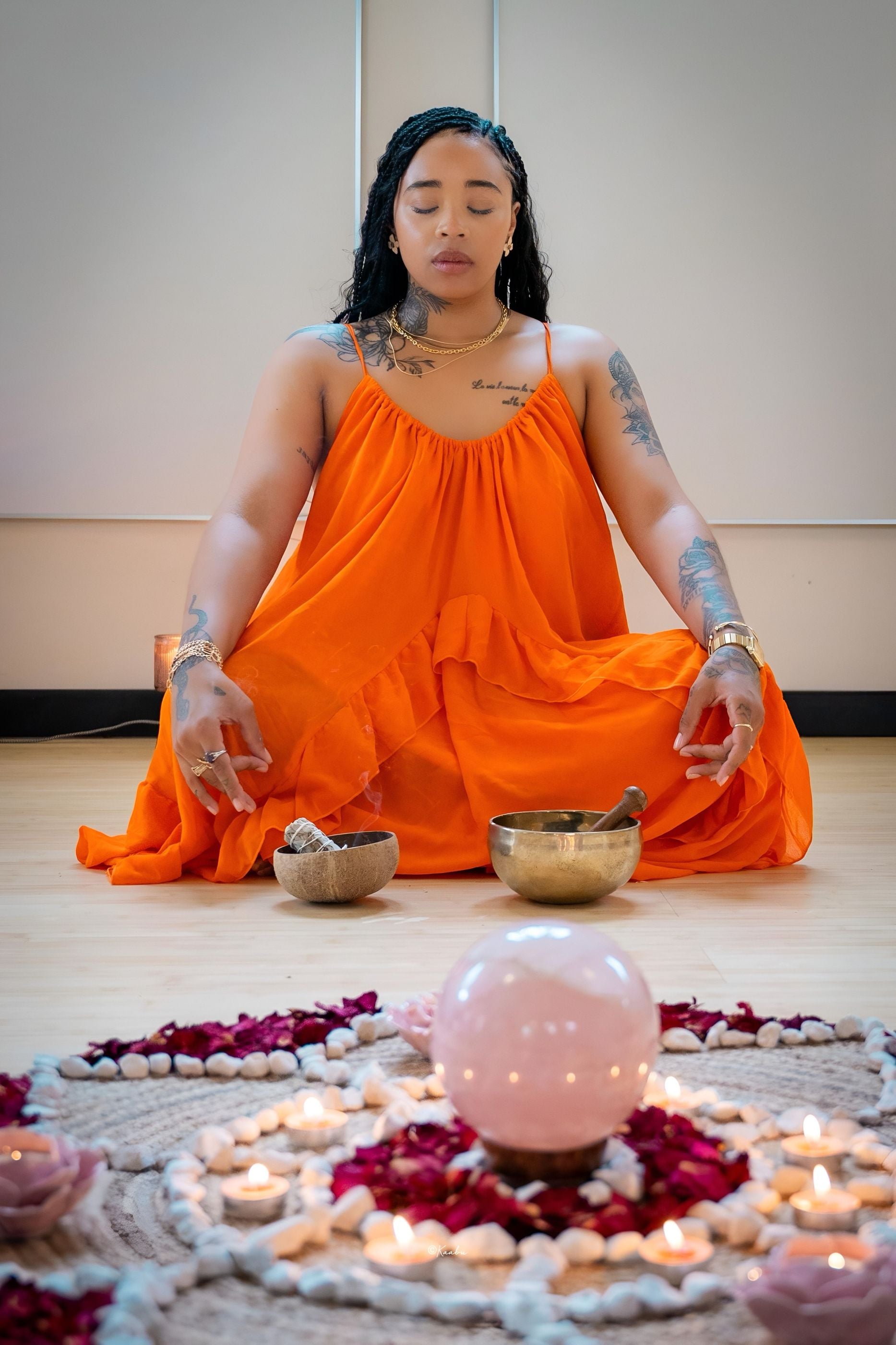 Océane médite en tailleur, en robe orange, derrière un mandala de pétales et une sphère de quartz rose